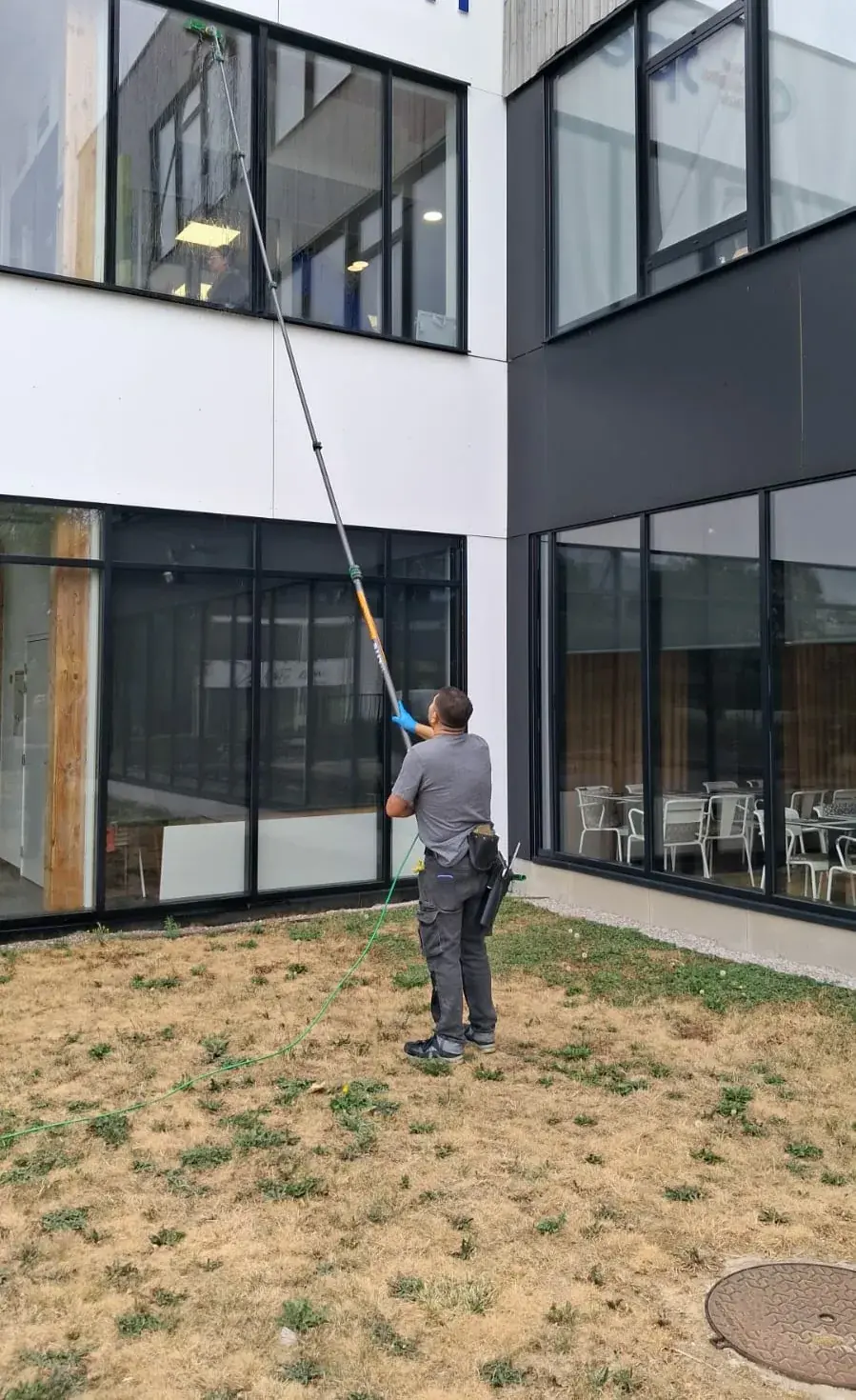 Agent réalisant le nettoyage de vitres extérieures en hauteur à l’aide d’une perche télescopique sur un bâtiment professionnel.