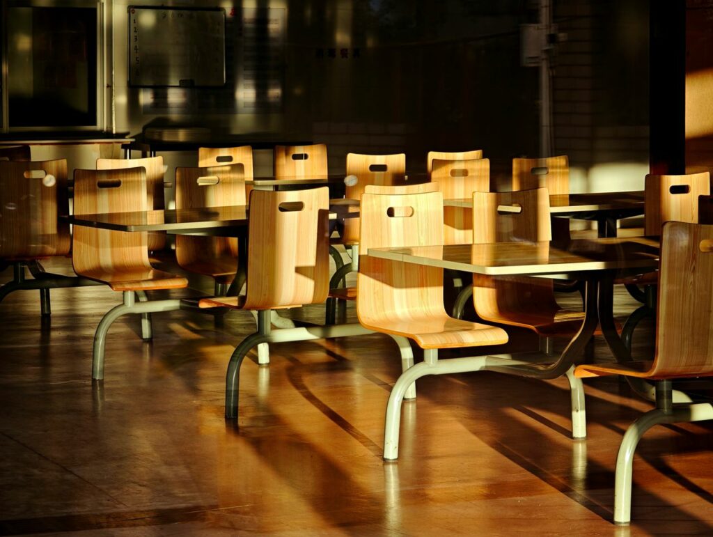 Empty wooden chairs and tables in a sunlit cafeteria setting, casting dramatic shadows.